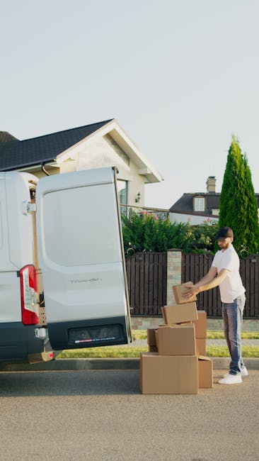 A man dressed in a beige knit beanie, a green padded vest over a dark blue jacket, and black gloves is seated on the edge of a driveway or pavement in front of an open van, with a focused and serious