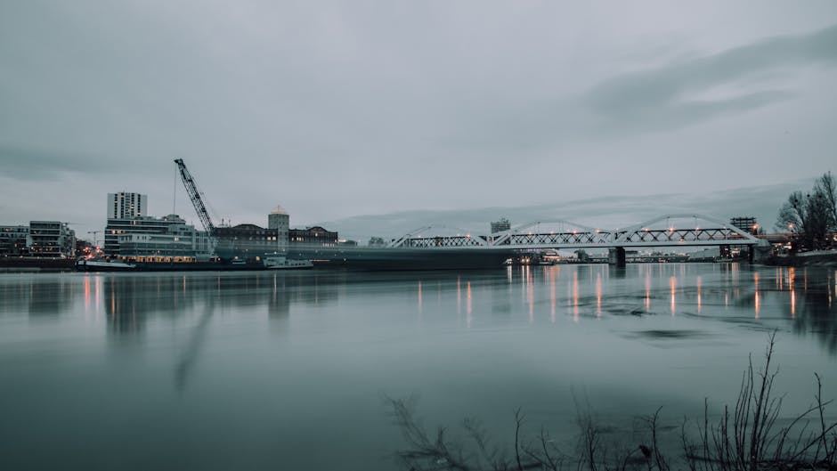 A wide view of the Kingston Bridge spanning over the River Thames during an overcast day, with the bridge illuminated by streetlights reflecting on the calm water surface. On the left side of the imag