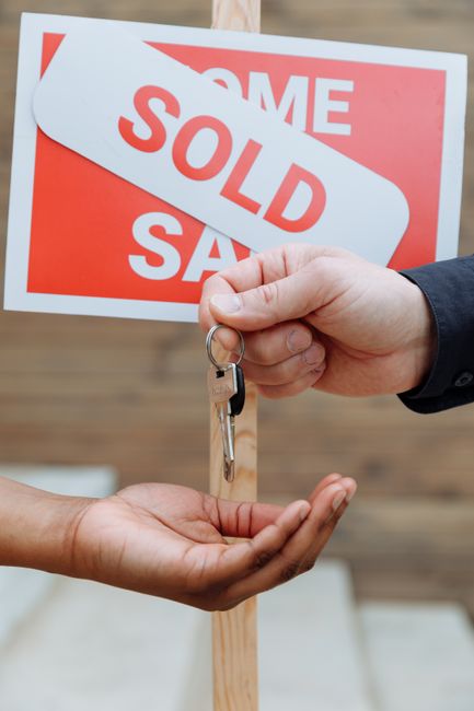 A person wearing a checkered coat is removing a white sticker with bold red letters reading 'SOLD' from a property for sale sign. The sign is attached to a wooden post and features red and white backg
